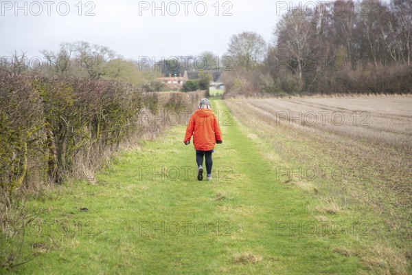 Person wearing bright orange coat walking along grassy path next to hedgerow and winter field, Sutton, Suffolk, England, UK