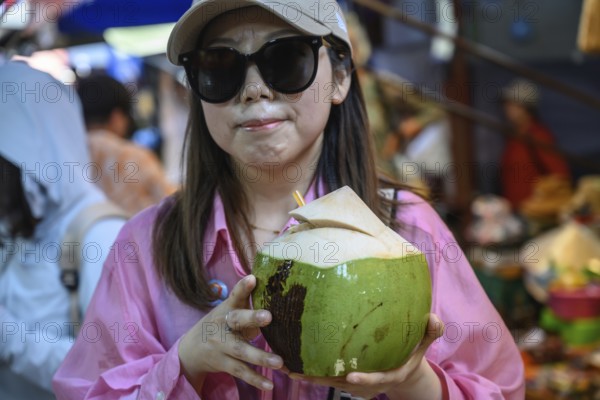 Mae Klong, Thailand. March 15th 2025. A smiling tourist with fresh coconut juice at the Mae Klong Railway market, in Samut Songkhram province of Thailand