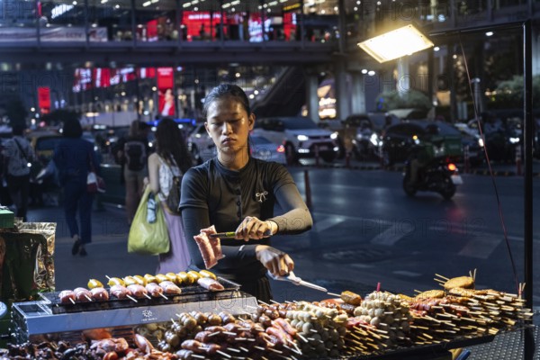 Bangkok, Thailand. March 26th 2025. A Thai woman cooks grilled meat at a street food stall at night in Siam Square a busy shopping district, Bangkok, Thailand, Asia
