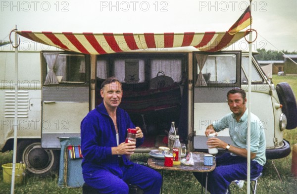 Two men having breakfast at campsite sitting outside a VW Volkswagen camper van vehicle, 1971