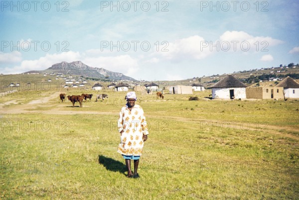 A local woman stands in the foreground of a rural Xhosa village (umuzi) in the Eastern Cape, South Africa. The village is nestled in a grassy, hilly landscape and features traditional circular huts known as rondavels