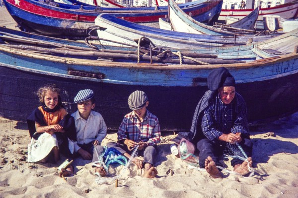 Fisherman and children repairing nets on beach by traditional fishing boats, Nazaré, Portugal, Europe 1968