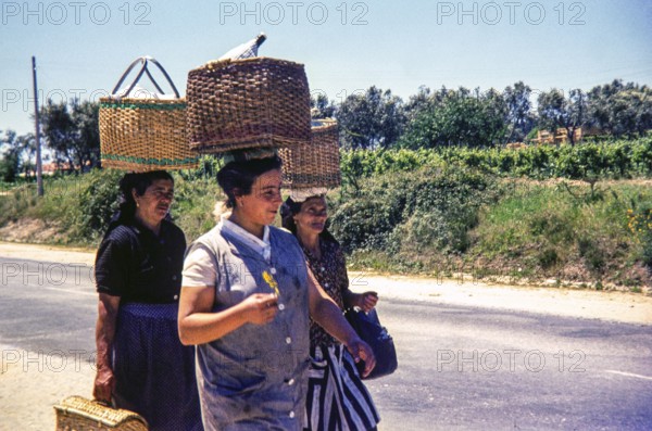Three women walking on street in rural area carrying wicker baskets on their head, Portugal, Europe 1968