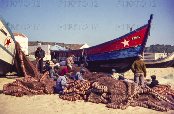 Fishermen repairing nets on beach by traditional fishing boats, Nazaré, Portugal, Europe 1968