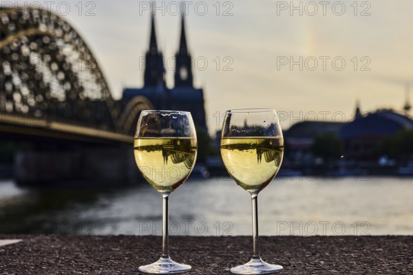 Wine glasses, wine, light refraction in filled glasses, concrete wall, Rhine river, Hohenzollern bridge, arched bridge, steel arches, Cologne Cathedral, double tower, general architecture, depth of focus, sharp foreground - background blurred, sunset, red and orange sky, cirrostratus clouds, Cologne, district-free city, North Rhine-Westphalia, Germany