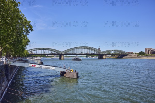 Rhine river, pedestrian and railway bridge Hohenzollern bridge, steel arches, general architecture, pier, gangway, Cologne-Düsseldorfer Deutsche Rheinschiffahrt GmbH, quay wall, water surface with small waves, trees, blue sky, cloudless, Leystapelwerft, Cologne, district-free city, North Rhine-Westphalia, Germany