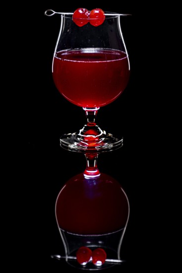 Red drink in a cognac glass with cherries against a dark background and reflection