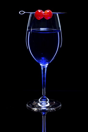 Blue drink in a wine glass with cherries, black background and reflection