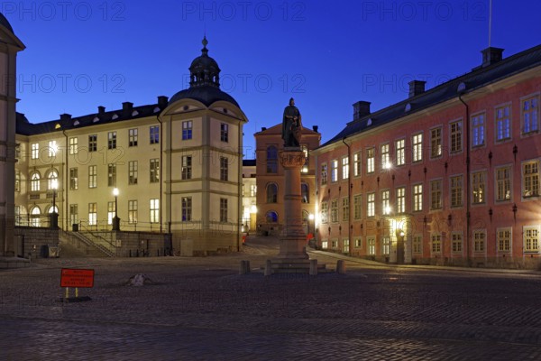 An illuminated town square with historic buildings and a memorial at dusk, Riddarholmen, Stockholm, Sweden