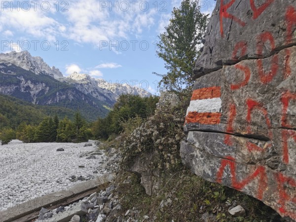 Rock with red and white markings, hiking trail, and mountain views under blue sky and clouds, Peaks of the Balkans, hiking in the Albanian Alps National Park, Albania