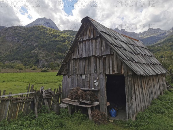 Rustic wooden cabin in a green meadow against an alpine mountain backdrop with cloudy sky, Peaks of the Balkans, hiking in the Albanian Alps National Park, Albania