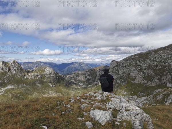 Person with backpack sitting on a rock and looking at a mountain panorama under cloudy sky, Peaks of the Balkans, hiking in the Albanian Alps National Park, Albania