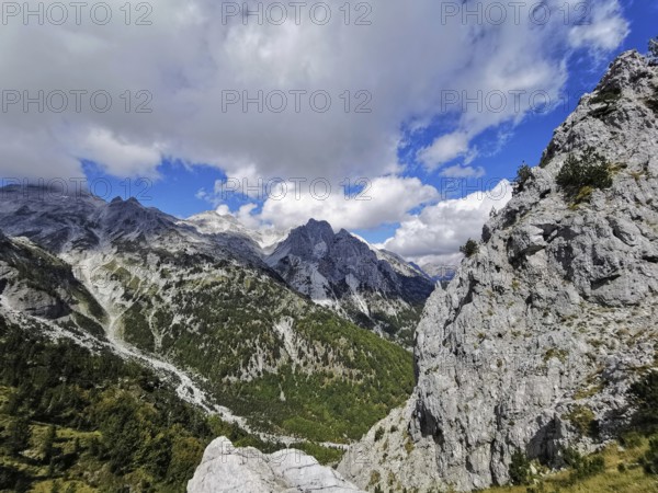 Rugged mountain landscape with clouds and blue sky, trees on rocky hillside, Peaks of the Balkans, hiking in the Albanian Alps National Park, Albania