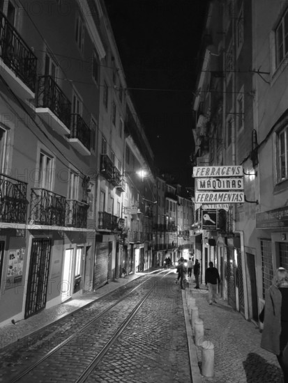 Nighttime street scene in a city with illuminated signs, tram tracks and people walking by, monochrome, Lisbon, Portugal
