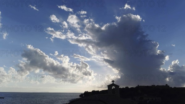 Dramatic sky with clouds and sunlight over sea with church silhouette in foreground, Himare, Albania
