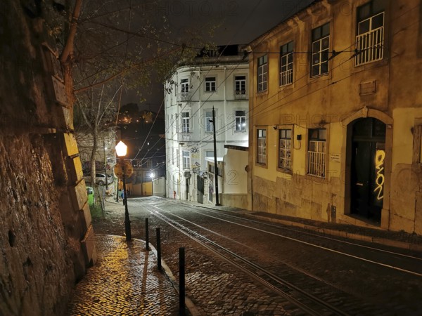 Night view of a picturesque street with cobblestones and buildings illuminated by lamps, tracks of the Elevador da Gloria funicular, Lisbon, Portugal