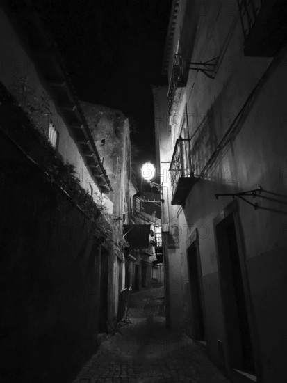 A narrow alley at night with illuminated alleyway lamp between tall, dark buildings, monochrome, atmospheric, Lisbon, Portugal
