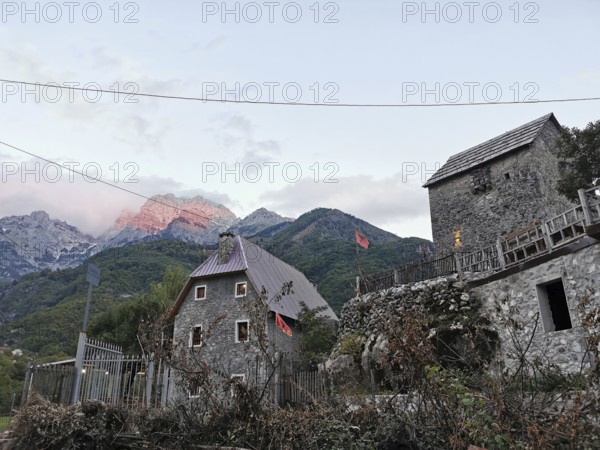 Traditional village with stone houses and Albanian flags against a mountain backdrop, mountain glow, alpine glow in the background, Peaks of the Balkans, hiking in the Albanian Alps National Park, Albania