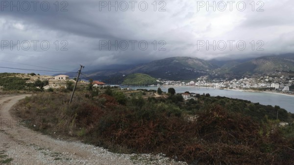Hilly coastal landscape with a path by the sea and a village on a mountainside under cloudy sky, Himare, Albania