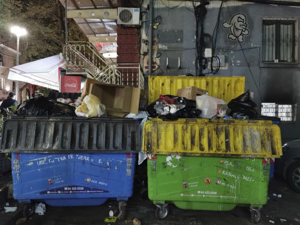 Two colored, overcrowded dumpsters on a street at night with graffiti on the wall, Himare, Albania