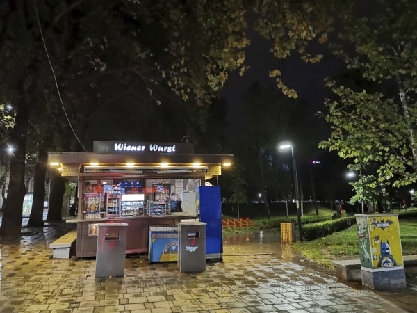 An illuminated sausage stand, Wiener Wurst, at night in a wet park, Tirana, Albania