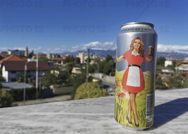 A colorful beer can with a woman in a red dress and a beer mug in her hand against a sunny urban background, Albania