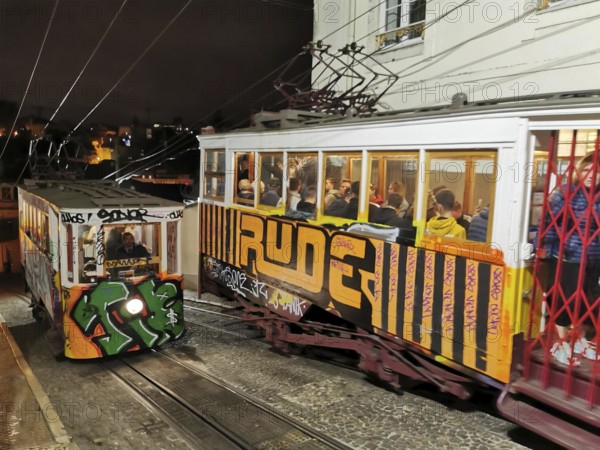 Two graffiti-painted funiculars travel illuminated through the night, Historic funicular, Elevador da Gloria with graffiti goes down a steep road, Lisbon, Portugal