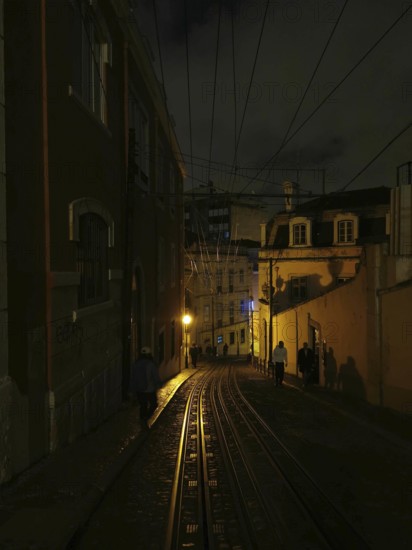 Rails of the Elevador da Gloria funicular, A dark alley at night with lantern light and shadows from passers-by, Lisbon, Portugal