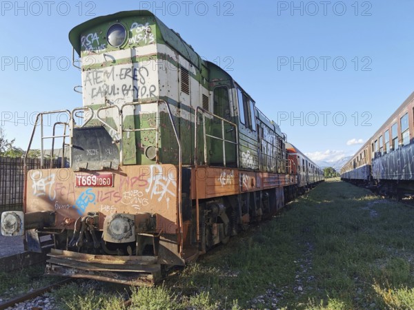 Graffiti-painted locomotive on rails, other abandoned trains next to it, Albania