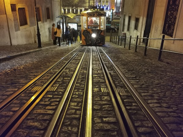 Front view from track perspective, graffiti-painted funicular runs illuminated through the night, historic funicular, Elevador da Gloria with graffiti goes up a steep road, Lisbon, Portugal
