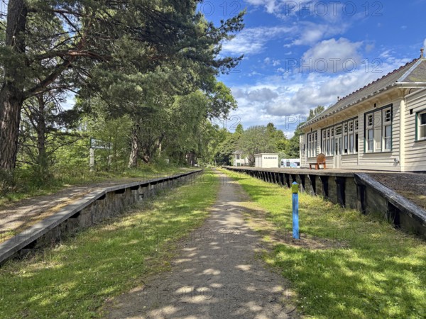 Abandoned station area with wooden buildings and tracks next to a wooded path under blue sky, railway track turned cycle path, Spey River, Aberdeenshire, Scotland, United Kingdom