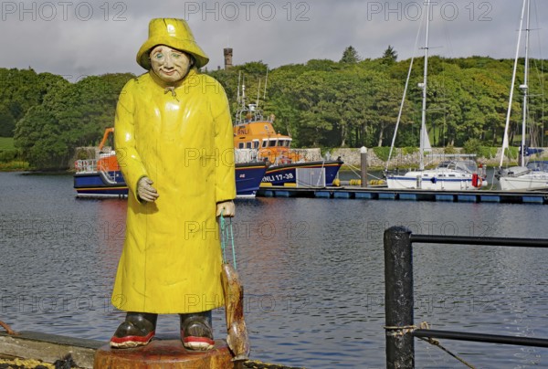 Statue of a fisherman in yellow raincoat at harbour in front of anchored boats with green hills in the background, Stornoway, Hebrides, Scotland, Great Britain