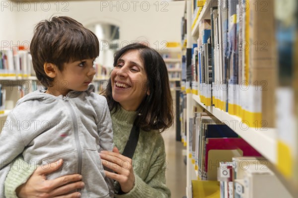 Mother holding her young child, both smiling and interacting happily while standing by shelves filled with books in a brightly lit public library, sharing a moment of learning and discovery
