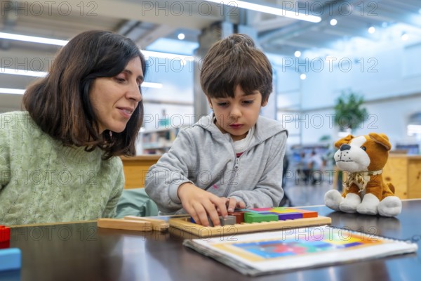 Teacher and young boy engaging in educational play, carefully fitting colorful geometric blocks into a wooden puzzle, fostering concentration and early learning skills in a bright library setting