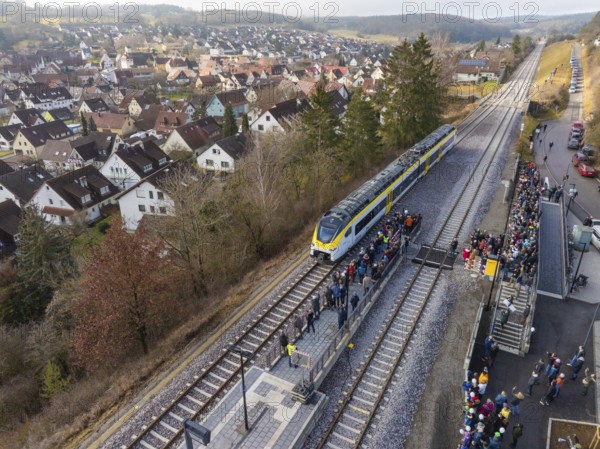 A train stops at a rural train station with surrounding people. The surrounding area shows a village and hills, opening of the Hermann Hesse Railway, Calw, Germany