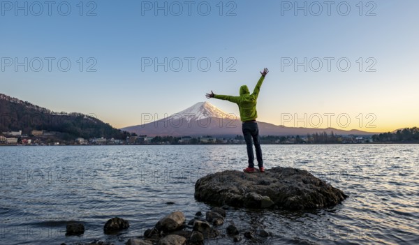 Young man stretching his arms in the air, standing on rocks in Lake Kawaguchi, view of Mount Fuji volcano at sunset, Yamanashi Prefecture, Japan