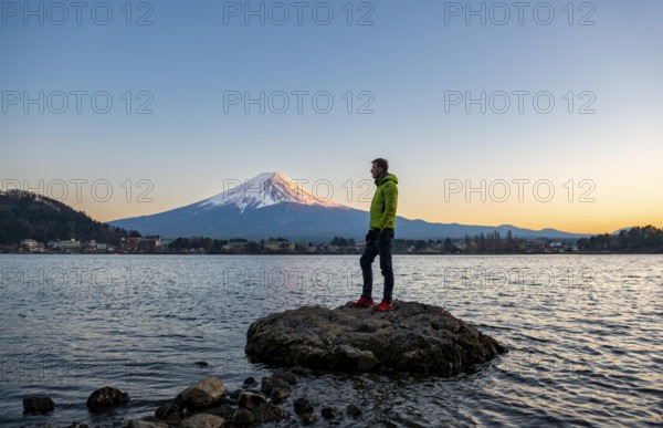 Young man standing on rocks in Lake Kawaguchi, view of Mount Fuji volcano at sunset, Yamanashi Prefecture, Japan