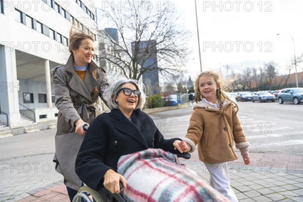 Family members, including a young girl and her mother, are pushing an elderly grandmother in a wheelchair outdoors, enjoying a cheerful moment together while leaving a building