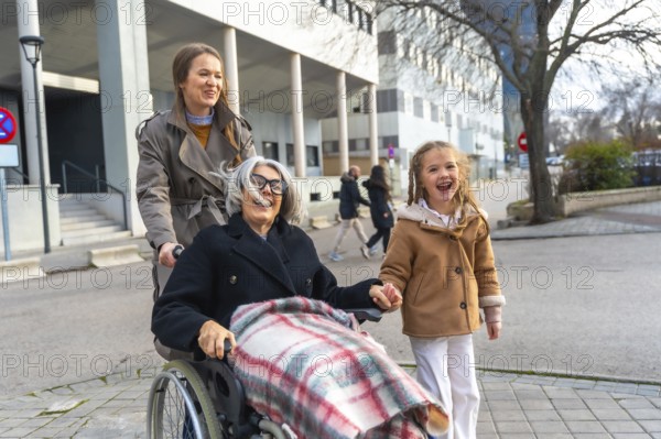 Three generations walking outdoors, smiling grandmother in wheelchair pushed by her daughter while granddaughter holds her hand, sharing joy, care and family connection
