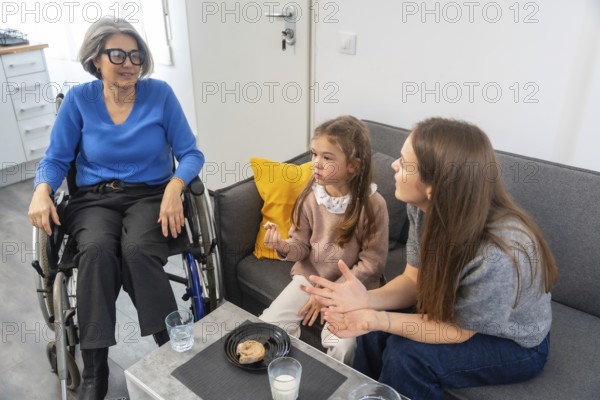 Granddaughter and mother enjoying a family visit with elderly grandmother sitting in a wheelchair, sharing a moment of connection and care in a modern living room setting