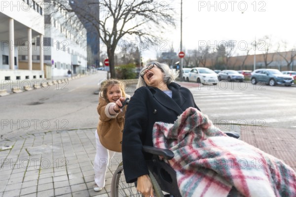 Granddaughter pushing her joyful grandmother in a wheelchair during an outdoor walk on a sunny day, sharing a happy moment of family connection and care