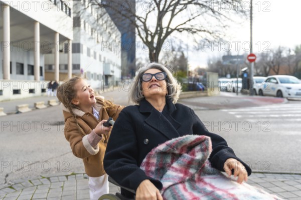 Happy granddaughter pushing senior grandmother in a wheelchair, enjoying a sunny day outdoors and fresh air, expressing love and intergenerational bond during a visit to the nursing home