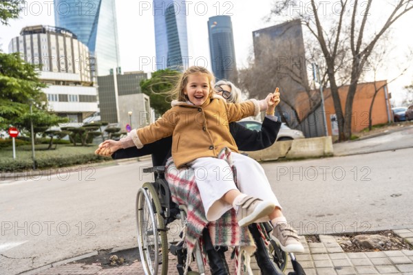 Happy little girl laughing and spreading arms, sitting on an older woman's lap in a wheelchair, enjoying a city park walk with modern buildings in the background