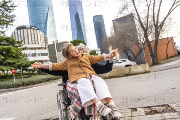 Happy granddaughter sitting on grandparent's lap in a wheelchair, holding hands and laughing with arms outstretched while enjoying a family moment outdoors with city skyscrapers in the background