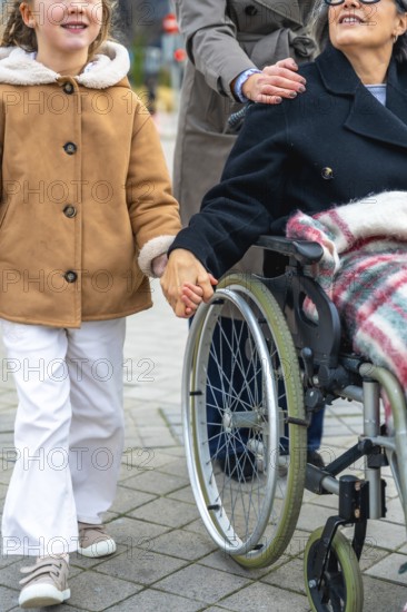 Granddaughter walking alongside her grandmother in a wheelchair, holding hands, with a woman pushing the wheelchair, representing family support and care for elderly people