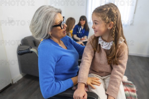 Grandma in a wheelchair interacting happily with her granddaughter, sharing a warm moment during a family visit at a facility with healthcare professionals present in the background