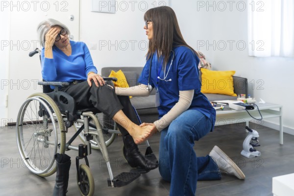 Medical professional providing physical therapy and attentive care to an elderly patient experiencing discomfort in her leg while sitting in a wheelchair at home