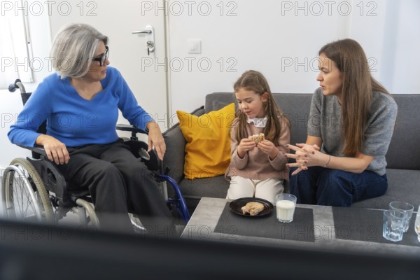 Grandmother in a wheelchair interacting with her family, including a young girl eating a snack and a woman speaking, sitting together on a modern sofa in a bright home setting