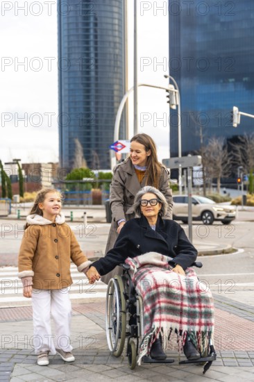 Three generations family, including little girl holding hands with her grandmother in a wheelchair, enjoying an outdoor walk with daughter pushing grandmother in cityscape