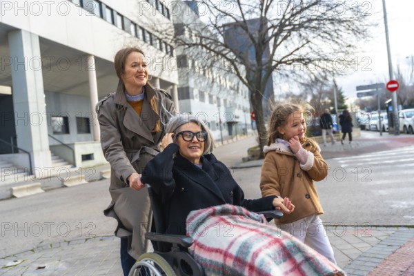 Three generation outing with adult daughter and little girl pushing smiling grandmother in wheelchair down a sunny urban sidewalk, enjoying supportive, caring family time outdoors